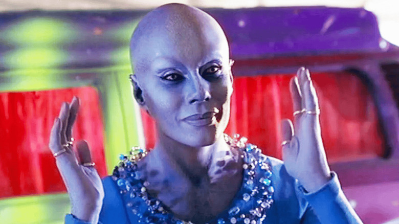 Pa'U Zhaan holds up her blue hands while wearing gold rings and a blue dress in a red room in "Farscape"