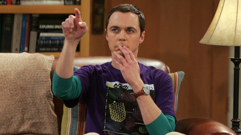 Sheldon sitting in his spot on the couch in "The Big Bang Theory" while covering his mouth with one hand and pointing with another