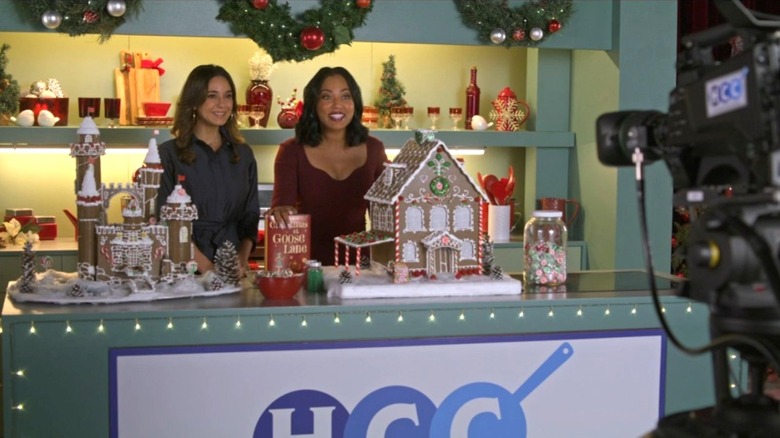 Joy and Liza stand behind gingerbread houses on a TV set in Joy to the World (2025)