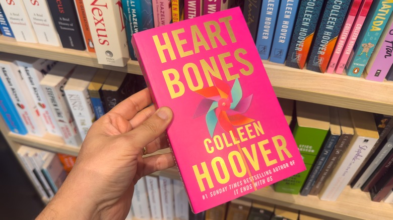 Someone holding Colleen Hoover's "Heart Bones" in front of a bookshelf