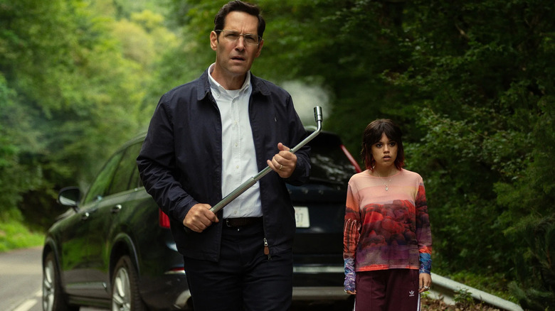 Elliot holding a tire iron and approaching something off-screen as his daughter stands by watching, both are flanked by a black SUV and a forest road