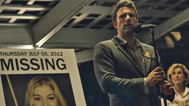 Nick speaking into a microphone in front of a picture of missing Amy in Gone Girl