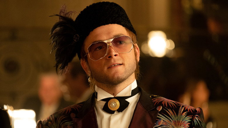 Elton John sitting down in feathered hat and patterned jacket in Rocketman (2019)