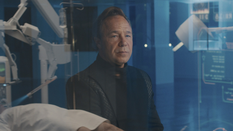 Elias Mannix sitting behind a hologram in "Bodies" (2023)