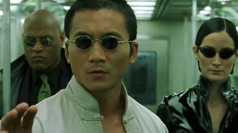Seraph wears small black sunglasses over a white shirt while Morpheus and Trinity stand behind him in "The Matrix Reloaded"