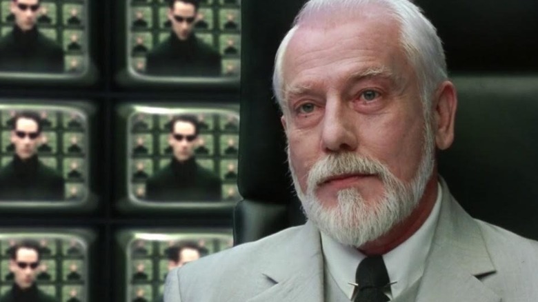 The Architect wears a white suit with black tie while seated in a leather chair in front of a wall of surveillance monitors showing images of Neo in "The Matrix Reloaded"