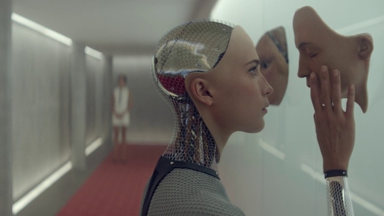 Ava touching an artificial face mounted on the wall in front of her with a woman standing in the blurred foreground