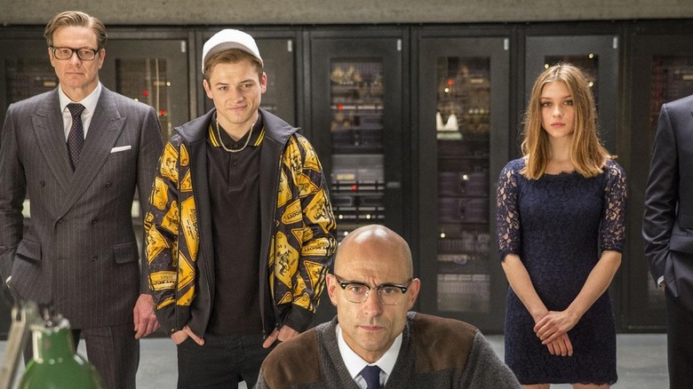 Galahad (Colin Firth), Eggsy (Taron Egerton), and Roxy/Lancelot standing behind Merlin (Mark Strong) as he works on the computer