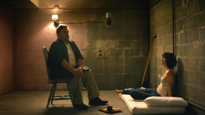 Michelle sitting on a mattress on the floor and looking up at Howard sitting on a chair in 10 Cloverfield Lane