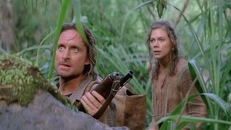 Jack hides behind a rock with a rifle in the jungle while Joanne looks concerned behind him in Romancing the Stone (1984)