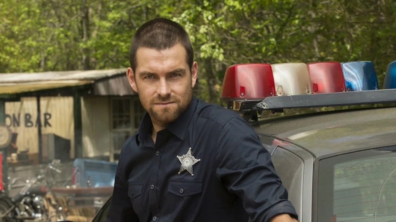 The ex-con stands by a cop car in the woods in Banshee