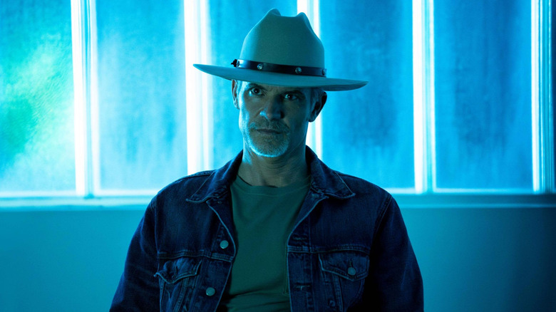 Raylan sits at in front of a window lit with neon blue in Justified: City Primeval