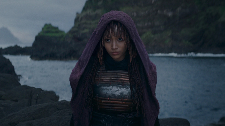 Mae Aniseya wears a purple cloak and hood over her braided hair while standing on a rock amidst the sea in "The Acolyte"