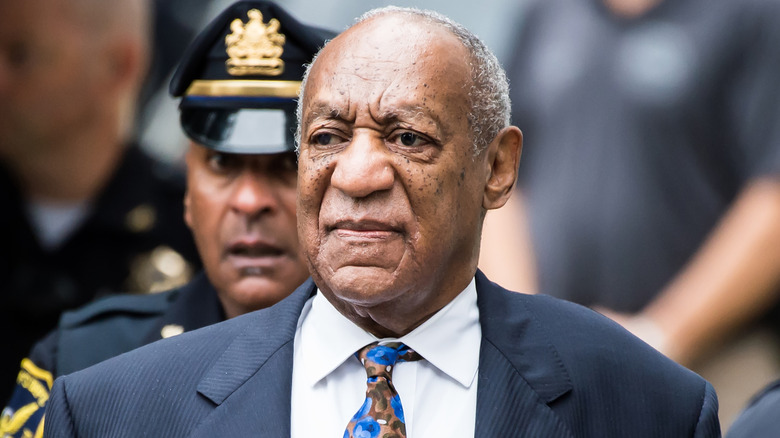 Bill Cosby on the street walking in front of a police officer, dressed for court