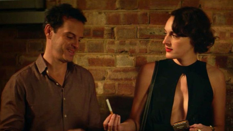 Fleabag, wearing a black cocktail dress with plunging cleavage, offering a cigarette to the Hot Priest as they stand in front of a brick wall