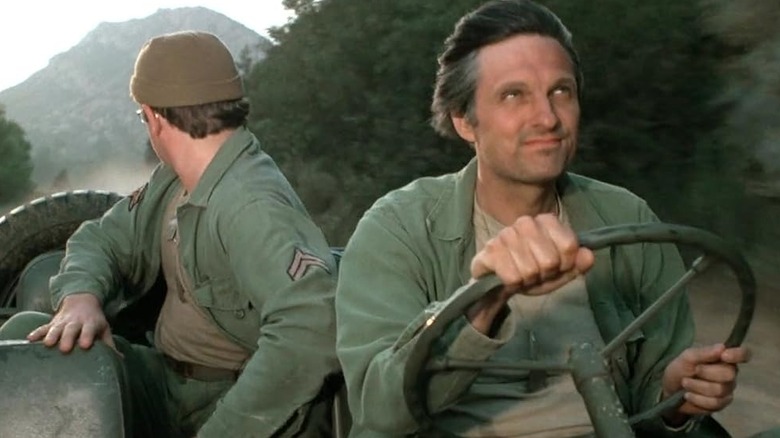 Hawkeye driving a military jeep with Radar in the backseat, looking over his shoulder