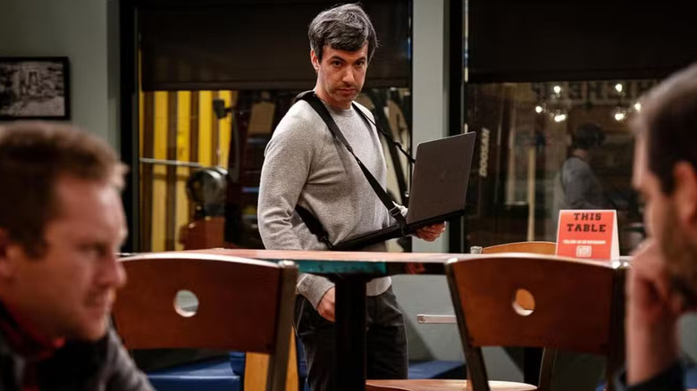 Nathan Fielder observing from the foreground and taking notes on a harnessed laptop as two people talk several feet away