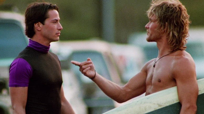Bodhi holding his surfboard and pointing at Johnny Utah in Point Break