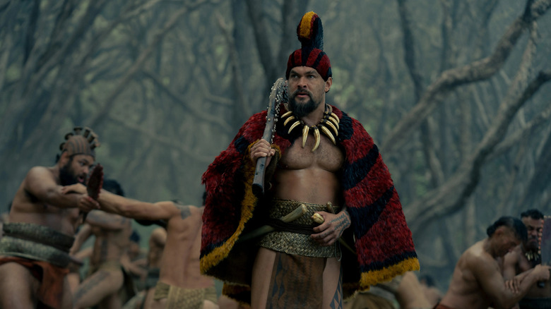 Ka'iana holds a club as he stands amongst his troopers while wearing a red and black cloak with a helmet in "Chief of War"