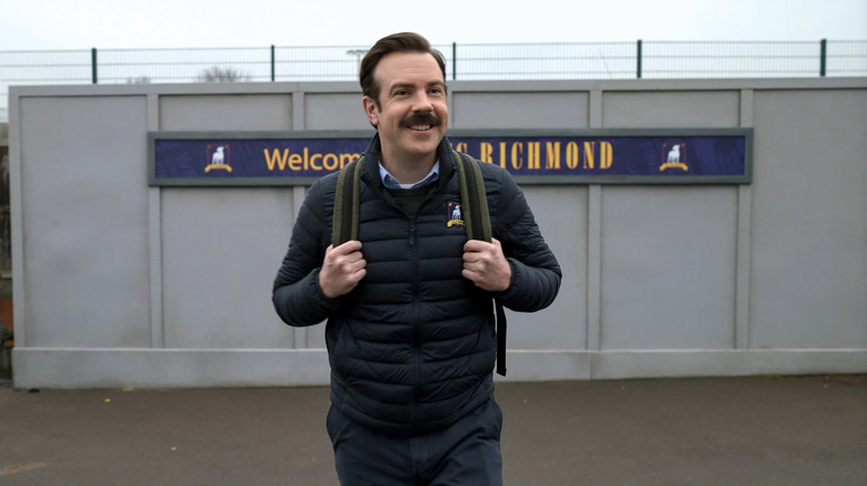 Ted Lasso holds his backpack with a big smile on his face while standing in front of a sign welcoming him to Richmond in "Ted Lasso"