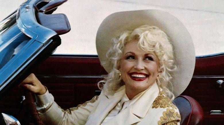 Mona driving a car and wearing a hat grinning in The Best Little Whorehouse in Texas