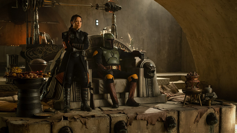 Boba Fett sits on his throne in his green armor while Fennec Shand stands beside him with her arms crossed in "The Book of Boba Fett"