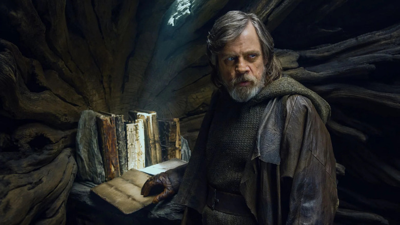 Luke Skywalker wears dirty brown clothes while standing inside a tree holding the ancient Jedi texts in 