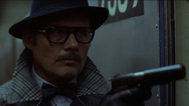 Mr. blue holds up a gun while wearing large sunglasses and a hat in "Taking of Pelham One Two Three"