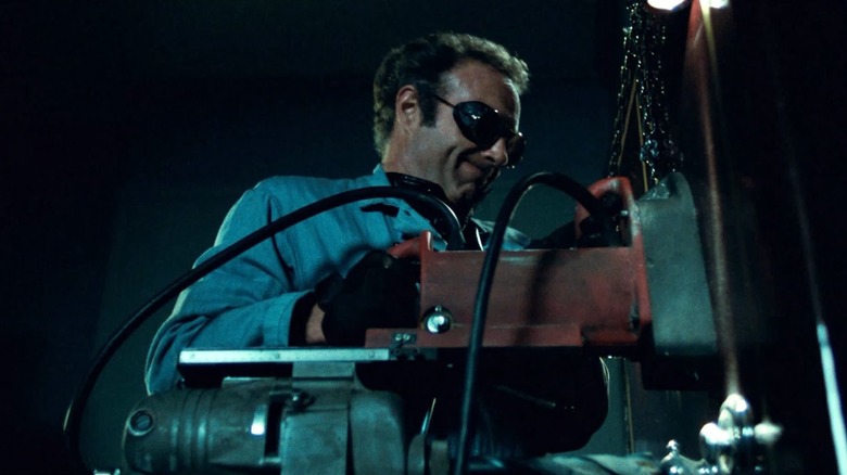 A man in a jumpsuit holds a large mechnical piece of hardware while wearing goggles in "Thief"