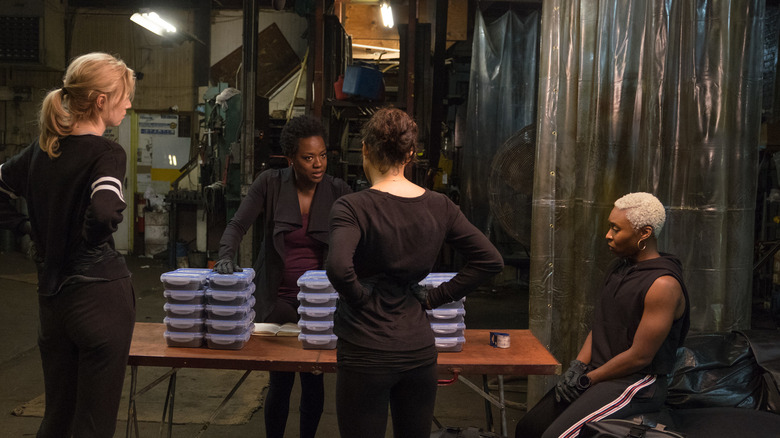 Four women stand around a table with stacks of items in front of them, all wearing black, in "Widows"