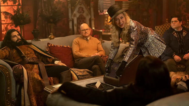 Nandor, Colin Robinson, and Guillermo siting on dark furniture in a macabre-looking room while The Guide (Kristen Schaal) prepares equipment in front of them