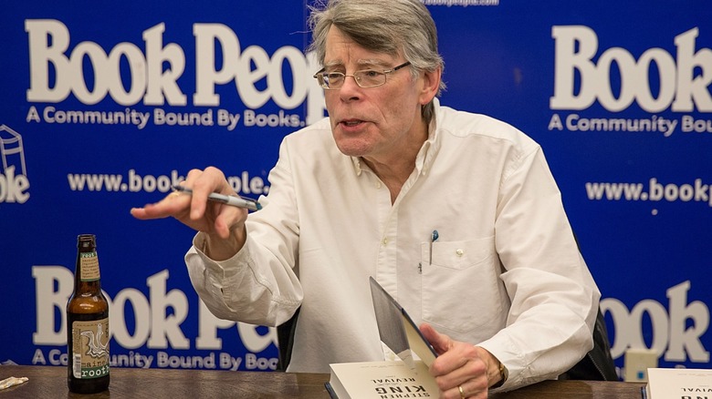 Stephen King at a book signing, an open book and a root beer on the table in front of him
