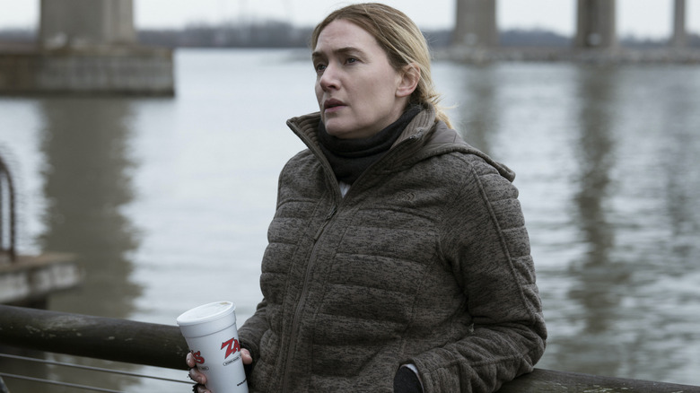 Mare by the river holding a coffee on Mare of Easttown