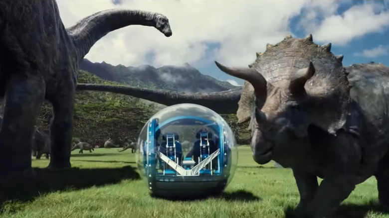 A tour vehicle passes by a brontosaurus and a triceratops in Jurassic World