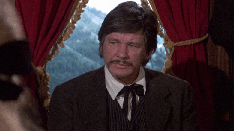 John Deakin standing in front of a window in "Breakheart Pass"
