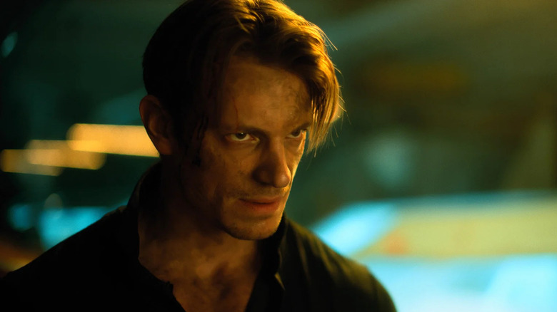 Kovacs sends a smoldering glare while standing in a dark space lit with dark yellow light