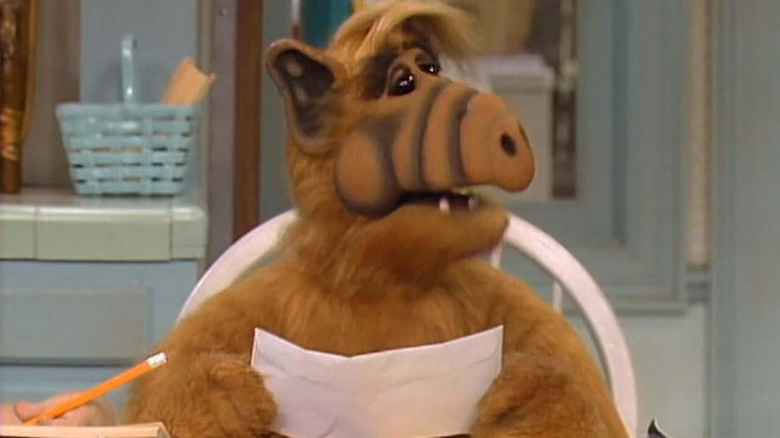 Gordon Shumway aka ALF sits at a table holding a piece of paper in ALF.