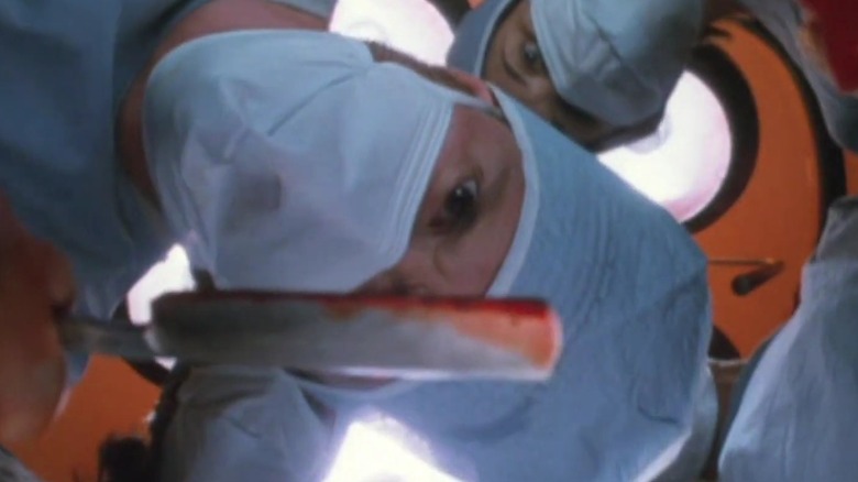 Masked-up surgeons leaning down toward the camera, holding up a bloody switchblade, in Kolobos