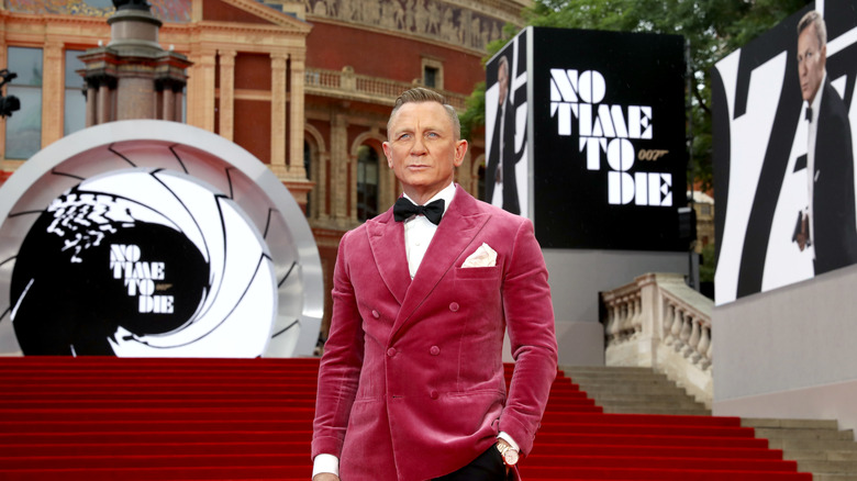 Daniel Craig in a bright mauve suit standing among signage for No Time to Die