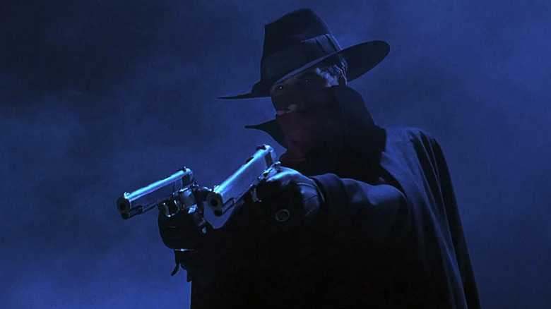The Shadow dressed in his iconic jacket, hat, and scarf, aiming with his dual pistols