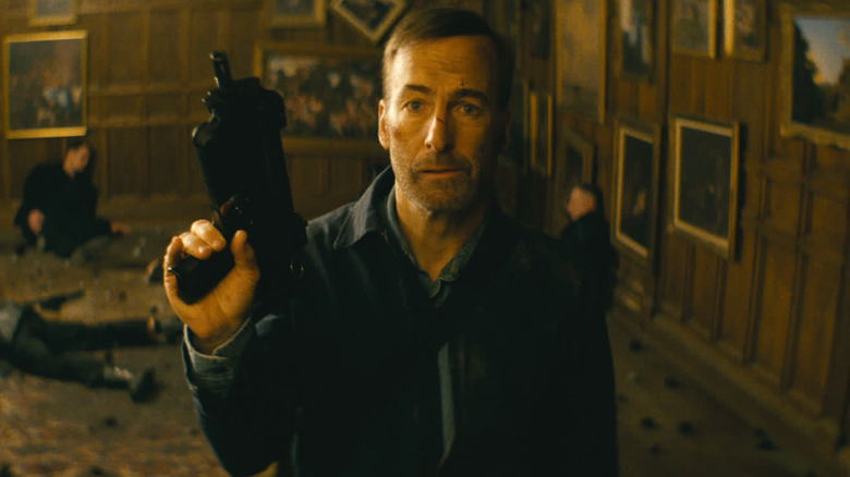 Hutch holding up a gun with bodies behind him in "Nobody" (2021)