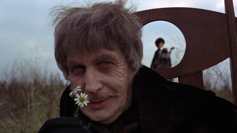A smiling Dr. Phibes holds flowers to his nose