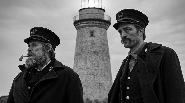 The two leads of The Lighthouse (2019) standing in front of the movie's titular location