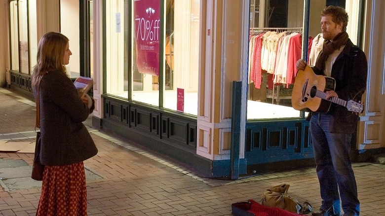 Girl faces Guy as he stands with his guitar on the sidewalk in "Once"