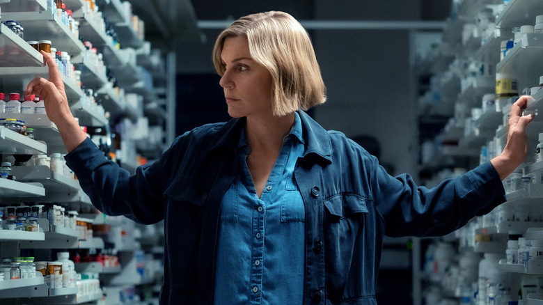 Carol walking through a narrow pharmacy aisle, holding onto shelves while scanning a plethora of pill bottles