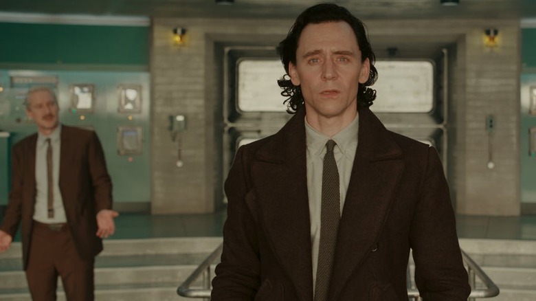 Loki standing with Mobius in the TVA, wearing a business suit and staring in the direction of the camera