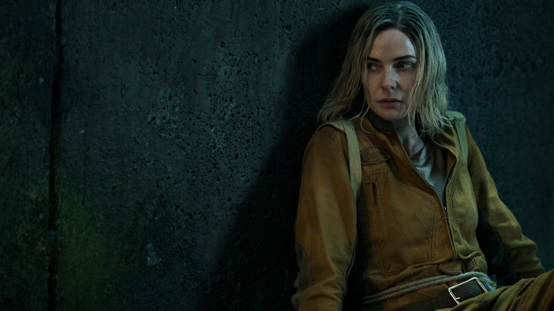 Juliette sitting against a dark, concrete wall dressed in an engineering jumpsuit and looking to the side