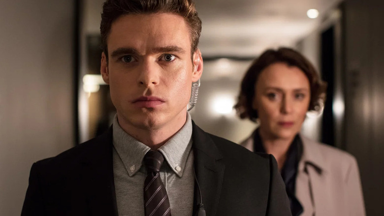 David stands in a hallway looking focused, while Julia stands behind him calmly.
