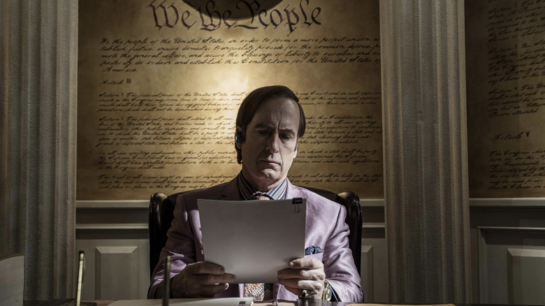 Saul Goodman sitting in office in front of constitution on wall