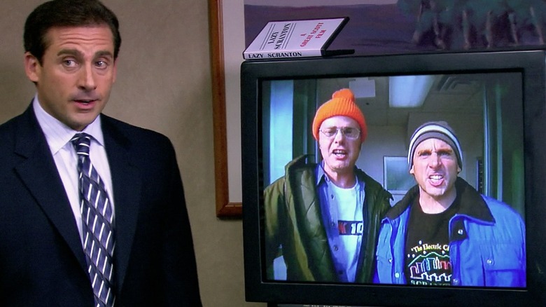 Michael Scott standing in the Dunder Mifflin conference room next to a TV showing his "Lazy Scranton" music video